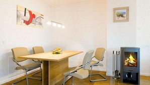 A modern dining area with a wooden table, four chairs, a fruit bowl, a wall-mounted light, and a wood-burning stove. - Modernes, Strandnahes Apartment mit Großem Balkon, Fußbodenheizung u. Kaminofen (Ostseebad Binz)