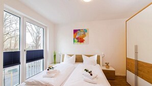 A hotel room with two beds, a large window, a colorful artwork on the wall, and a wooden floor. - Modernes, Strandnahes Apartment mit Großem Balkon, Fußbodenheizung u. Kaminofen (Ostseebad Binz)
