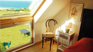 A room with a large window offering a view of the sea, a wooden chair, a small table with a lamp, and a bed with a red cover.