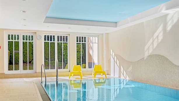 Una piscina interior con paredes de vidrio transparente, suelo de baldosas y sillas amarillas a su alrededor.