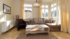 Una sala de estar con un sofá, una mesa de centro, una silla y ventanas amplias con cortinas.