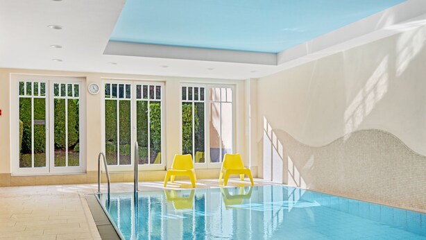Ein Hallenbad mit großen Glaswänden, gefliestem Boden und gelben Liegestühlen am Pool.