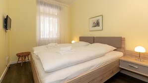 Ein ordentlich bezogenes Bett mit weißen Leinentüchern, ein Nachttisch mit Lampe, ein kleiner Korbstuhl und ein gerahmtes Bild an der Wand.