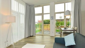 A modern living room with a sofa, a coffee table, and a dining area with a table and chairs. Large windows provide a view of the outdoors.