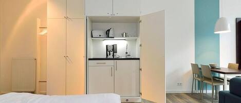 A compact hotel room with a bed, a kitchenette with a coffee machine, a dining table with chairs, and a view of the outside.