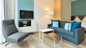 A modern hotel room with a grey armchair, a blue sofa with pillows, a small white table, a TV mounted on the wall, and a floor lamp.