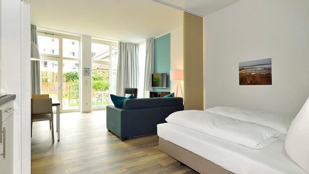 A modern hotel room with a large bed, a sofa, a TV, and a dining area with a table and chairs.