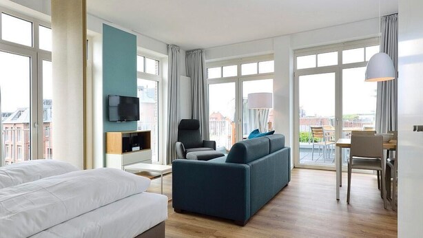 A modern hotel room with a large bed, a sofa, a TV, and a balcony with a table and chairs. - Tolles Ferienapartment am Inselstrand mit Meerblick, Balkon und Strandkorb! (Nordseeheilbad Wangerooge)