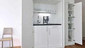 A compact kitchenette with a coffee maker, a chair, and open cabinets revealing kitchenware. - Tolles Ferienapartment am Inselstrand mit Meerblick, Balkon und Strandkorb! (Nordseeheilbad Wangerooge)
