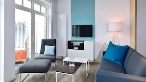 A modern living room with a sofa, armchair, and ottoman, a flat-screen TV mounted on a wall, and a window with curtains.