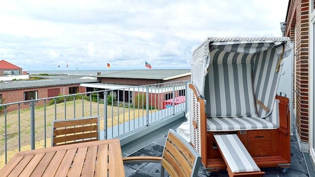 A balcony with a wooden table and chairs, a striped beach chair, and a view of the beach and buildings in the background.