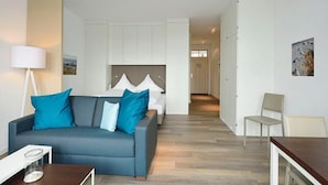 A modern hotel room with a grey sofa, a bed with a grey headboard, a white desk, and a chair.
