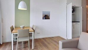 Un coin repas avec une table en bois, des chaises blanches, un luminaire suspendu blanc et un petit tableau encadré accroché au mur.