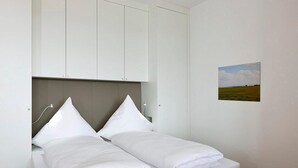 Un lit simple avec une literie blanche, une tête de lit et un petit tableau représentant un paysage accroché au mur.