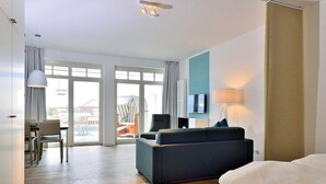 Ein modernes Hotelzimmer mit einer Couch, einem Fernseher, einem Essbereich mit Stühlen und einem großen Fenster.