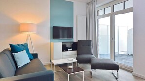 Una sala de estar moderna con sofá, sillón y reposapiés, un televisor de pantalla plana montado en la pared, una mesita y un amplio ventanal corredera que da a un espacio exterior.