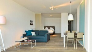 A modern living room with a sofa, a dining table with chairs, a bed, and a ceiling fan.