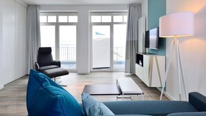 A modern living room with a sofa, armchair, and a TV mounted on the wall. There is a floor lamp and a window with curtains.