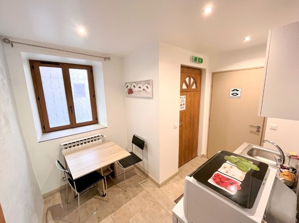 Une petite pièce bien éclairée, avec une table et des chaises en bois, une fenêtre avec des stores, une kitchenette équipée d’un évier et une porte portant un panneau indiquant la sortie.