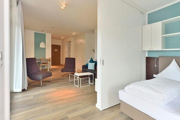 A modern hotel room with a bed, a desk, and a chair. There is a dining area in the background. - Hochwertiges Apartment an der Nordsee mit Sonniger, Großer Loggia in Südlage (Nordseeheilbad Wangerooge)