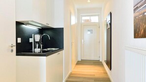 Un pasillo estrecho con un área de cocina a la izquierda, que incluye una encimera negra, una cafetera y un lavabo. El pasillo tiene piso de madera y paredes blancas.