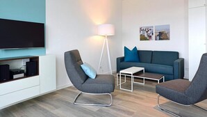 Una sala de estar moderna con un televisor de pantalla plana, dos sillones grises, un sofá azul, una mesa de centro blanca y una lámpara de pie.