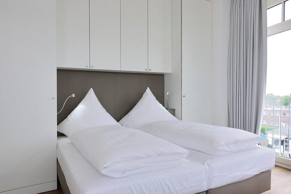 Una cama doble con ropa de cama blanca en una habitación de hotel con cabecera gris y paredes blancas.