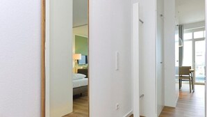 Un pasillo con piso de madera, paredes blancas y una puerta que da a un dormitorio con cama y mesita de noche.