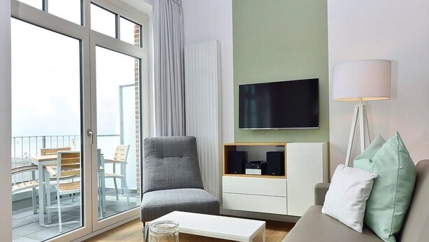 Una sala moderna con un sofá, una mesa de centro de vidrio, un sillón gris, un televisor en la pared y un balcón con mobiliario exterior.