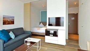 A modern hotel room with a grey sofa, a small white coffee table, a bed with white linens, a TV mounted on a white cabinet, and a wooden floor.