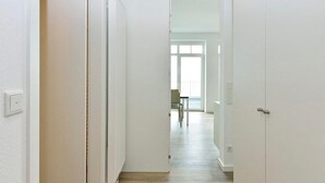 A narrow hallway with white walls and doors, leading to a room with a large window and a dining area visible in the background.