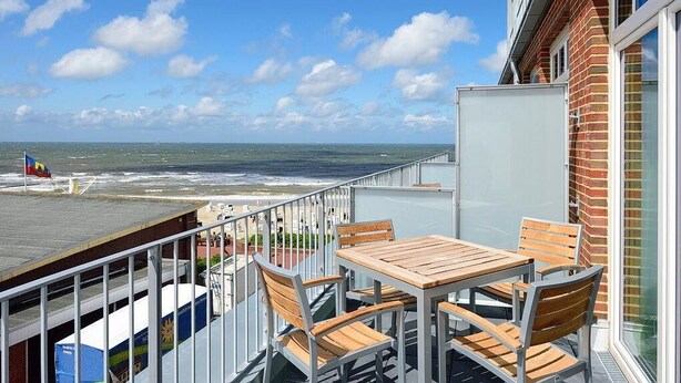 A balcony with a wooden table and chairs, overlooking a beach with a flag and ocean view. - Modernes Apartment an der See mit Westbalkon, Meerblick und Saunabereich im Haus (Nordseeheilbad Wangerooge)