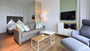 A modern living room with a grey sofa, a wooden coffee table, a grey armchair, a flat-screen TV mounted on the wall, and a white cabinet with speakers.