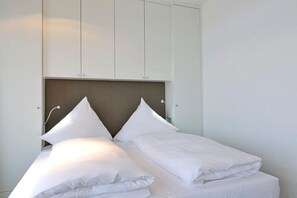 Uma cama com roupa de cama branca e cabeceira, um guarda-roupa branco acima e uma luminária fixada na parede.
