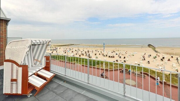 Ein Balkon mit Korbstuhl, Blick auf einen Strand mit Liegestühlen und einem gepflasterten Weg.