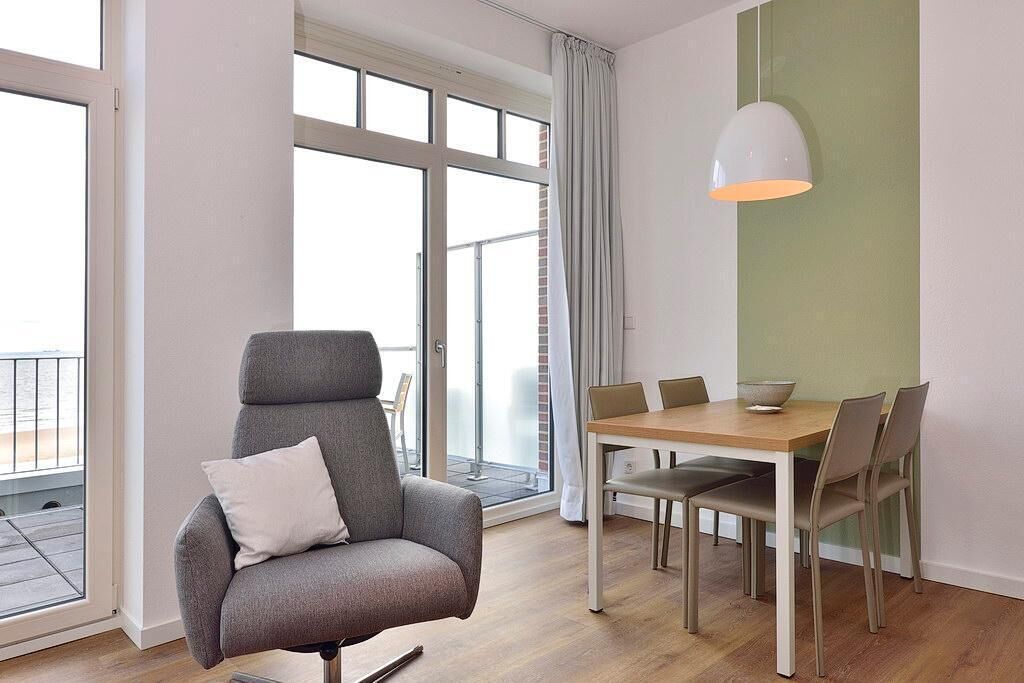 Un salon moderne avec un fauteuil gris, une table à manger en bois avec des chaises, une grande fenêtre avec des rideaux et un luminaire suspendu.