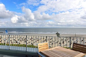 Un balcon avec une table et des chaises en bois, donnant sur une plage parsemée de nombreux parasols et ornée d’un drapeau.
