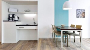 Una cocina moderna con armarios blancos, encimera negra y una cafetera. Hay una zona de comedor con una mesa de madera, dos sillas y una lámpara colgante.