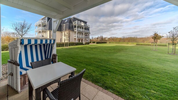 A covered patio area with a table and chairs, a striped outdoor seating area, and a view of a modern building across a well-maintained lawn.