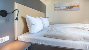 A neatly made bed with white linens and pillows, a bedside table with a lamp, and a painting of a beach scene on the wall.
