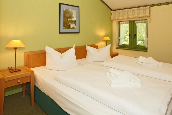 Habitación de hotel con dos camas, una mesita de noche con lámpara, un cuadro enmarcado en la pared y una ventana con cortinas.
