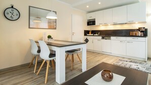 Eine moderne Küche mit weißen Schränken, einem Holztisch und Stühlen. An der Wand hängt eine Uhr und über dem Tisch befindet sich eine Pendelleuchte.