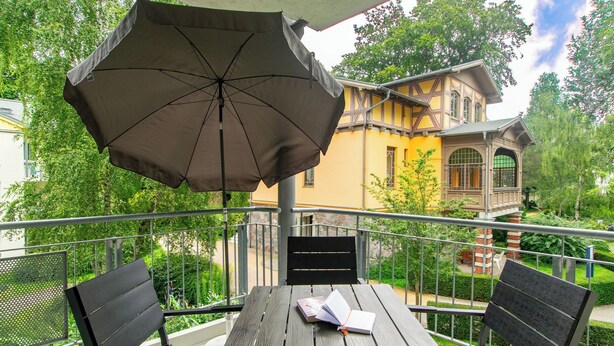 Ein Balkon mit Tisch, Stühlen und Sonnenschirm, mit Blick auf ein gelbes Gebäude mit Balkon.