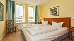 Habitación de hotel con dos camas, una silla de madera, una ventana con cortinas, un cuadro en la pared y una lámpara en la mesita de noche.