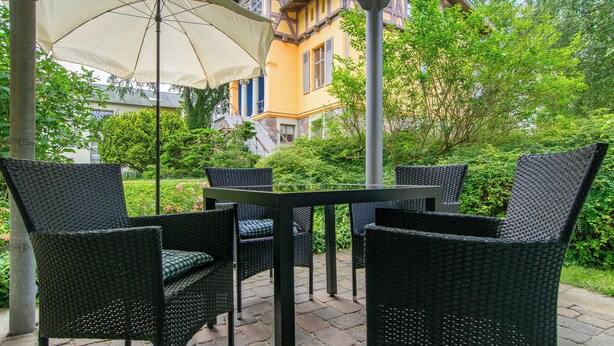 Un patio al aire libre con muebles de mimbre y una mesa, rodeado de vegetación y un edificio al fondo.