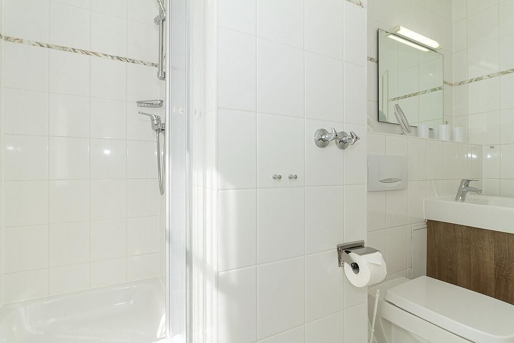 A modern bathroom with white tiles, a shower, a toilet, and a sink with a mirror above it.