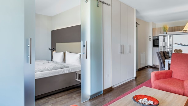 A modern room with a sliding door separating a bedroom and a living area. There is a bed with white bedding, a red chair, and a small table with a bowl of items.