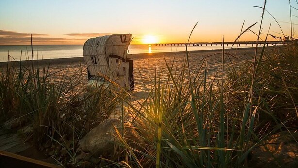 Una silla de playa en la orilla arenosa con un atardecer de fondo y un muelle que se adentra en el agua.