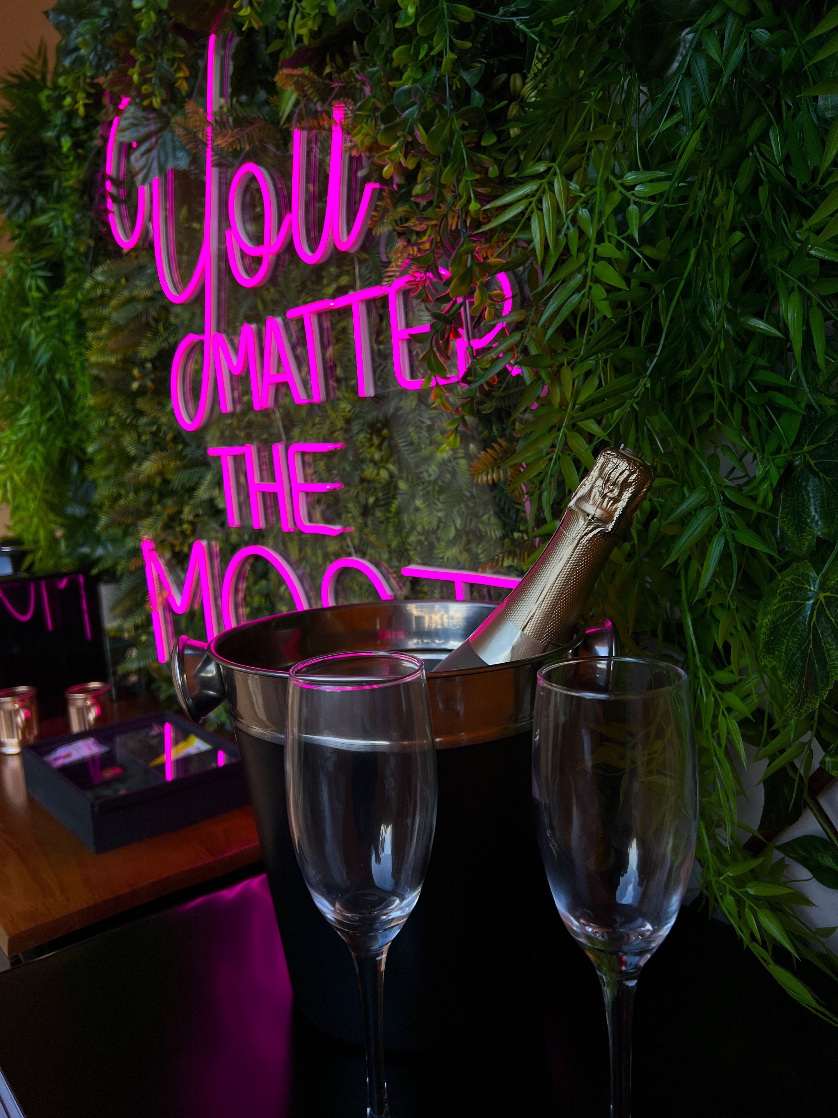 A bar area with a neon sign, a bottle of champagne, and two glasses on a wooden table.