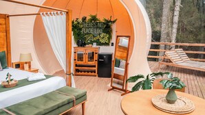 A cozy tented accommodation with a bed, a small table, a chair, and a view of the forest.